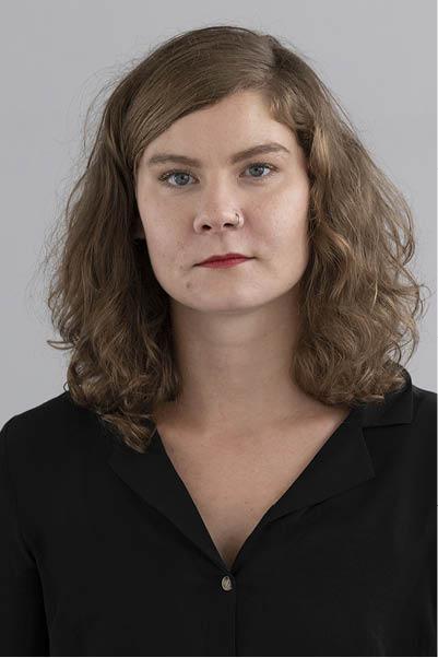 Oberkörper-Portrait einer jungen Frau mit braunem, lockigen Haar vor einem hellgrauen Hintergrund. Sie trägt roten Lippenstift und eine schwarze Bluse mit V-Ausschnitt. 