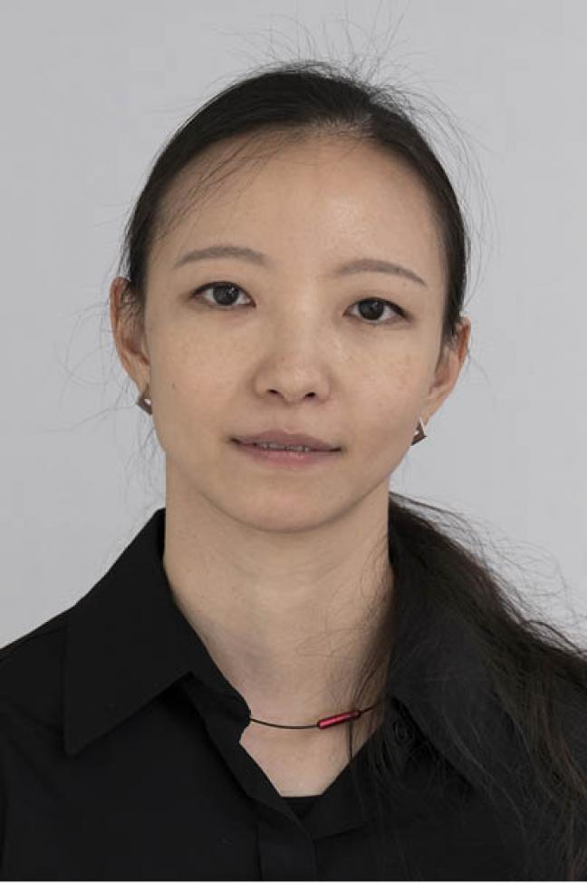 Oberkörper-Portrait einer Frau mit langem dunklen Haar in einem Pferdeschwanz vor einem hellgrauen Hintergrund. Sie trägt eine schwarze Bluse.