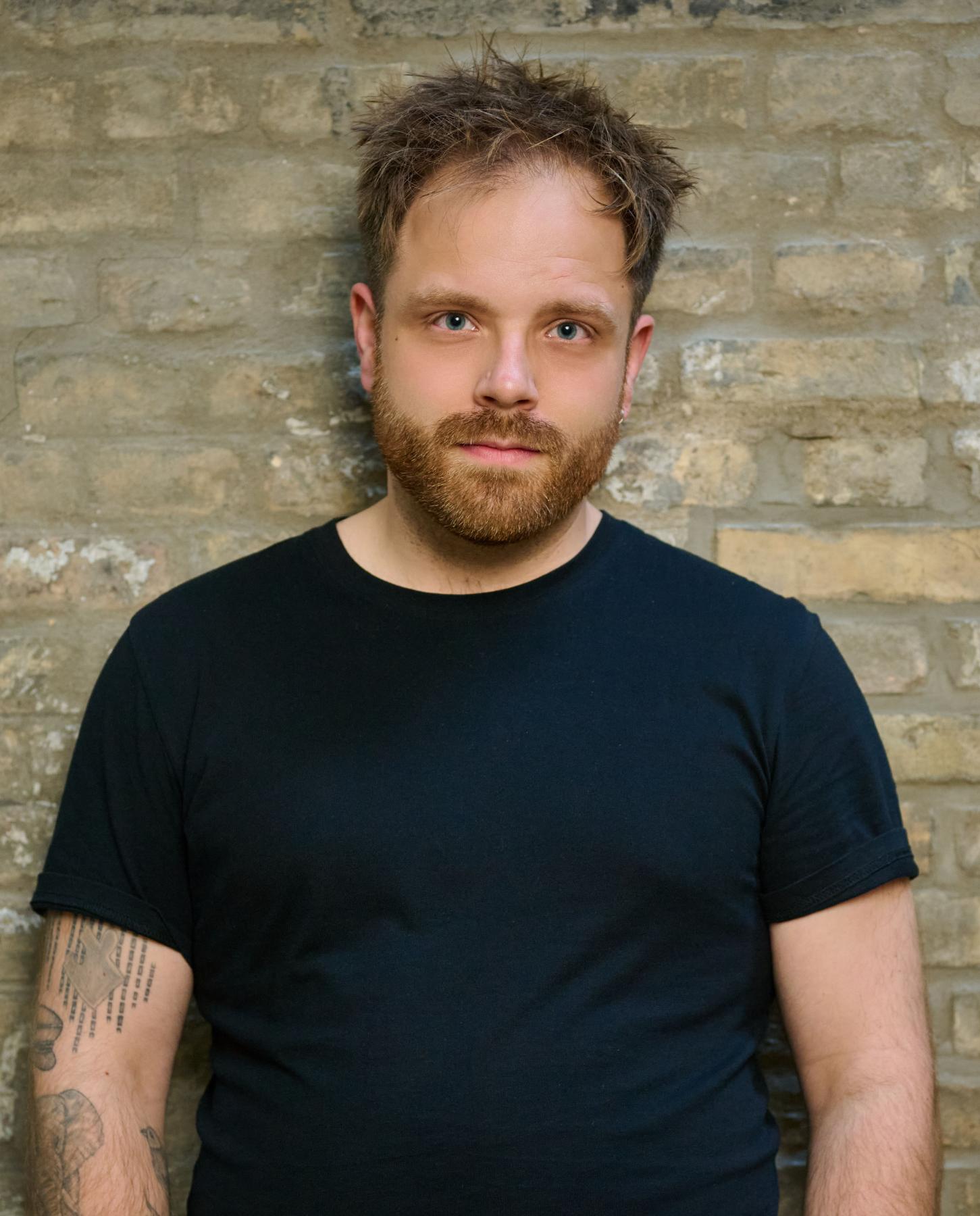 Portrait von Killian Prigge, ein junger Mann im schwarzen T-Shirt vor einer blassgelben Backsteinwand