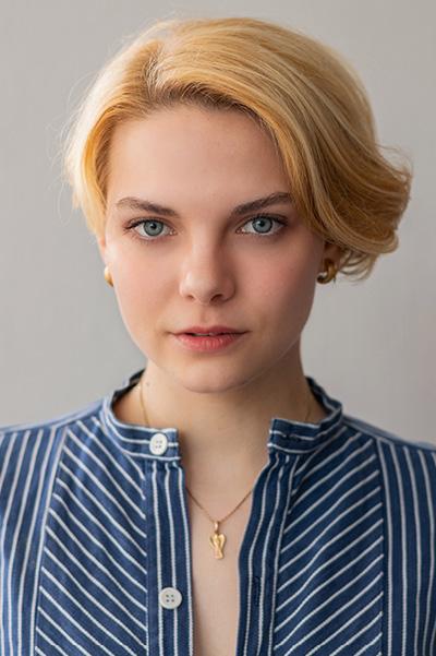 Oberkörper-Portrait einer jungen Frau mit kurzem, blonden Haar. Sie trägt ein blau-weiß gestreiftes Oberteil und steht vor einer hellen Wand. Dabei schaut sie mit einem interessierten Blick direkt in die Kamera.