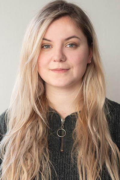 Oberkörper-Portrait einer jungen Frau mit langem, blonden Haar. Sie trägt einen dunklen Pullover und steht vor einem hellen Hintergrund. Dabei schaut sie mit einem Lächeln direkt in die Kamera.