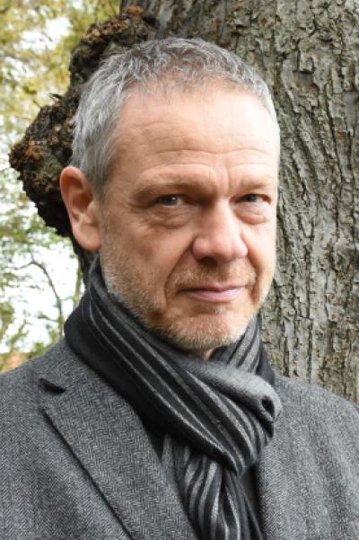 Ein Mann mit kurzem grauen Haar trägt einen grauen Mantel und einen grauen Schal. Er steht vor einem Baum und schaut mit einem neutralen Blick direkt in die Kamera.