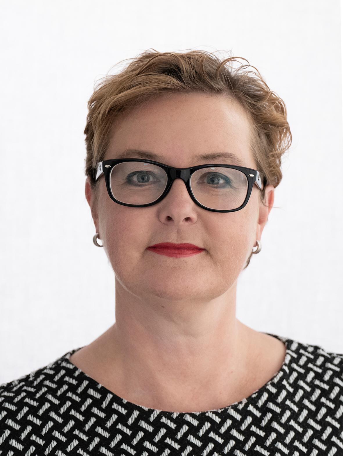 Portrait einer rotblonden Frau mit schwarzer Brille und schwarz-weißem Oberteil vor weißem Hintergrund. 