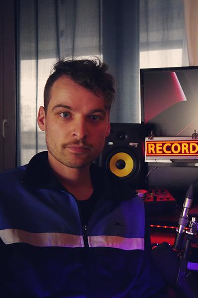 Oberkörper-Foto eines jungen Mannes mit kurzen dunklen Haaren und Oberlippenbart, der eine Trainingsjacke trägt. Er steht in einem Aufnahmestudio, rechts neben ihm ist ein Schild mit dem leuchtenden Wort RECORD zu sehen.