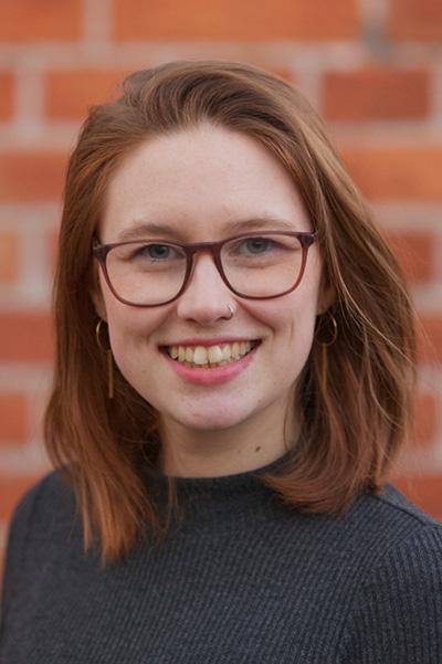 Oberkörper-Portrait einer jungen Frau mit rötlich-braunem Haar und einer roten Brille sowie einem Nasenpiercing vor einer roten Backsteinwand. Sie trägt einen blauen Pullover und lächelt in die Kamera.