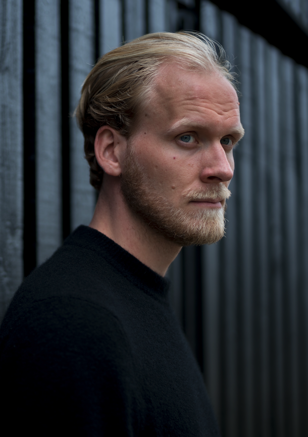 Ein blonder Mann steht vor einer Wand und schaut nach rechts.