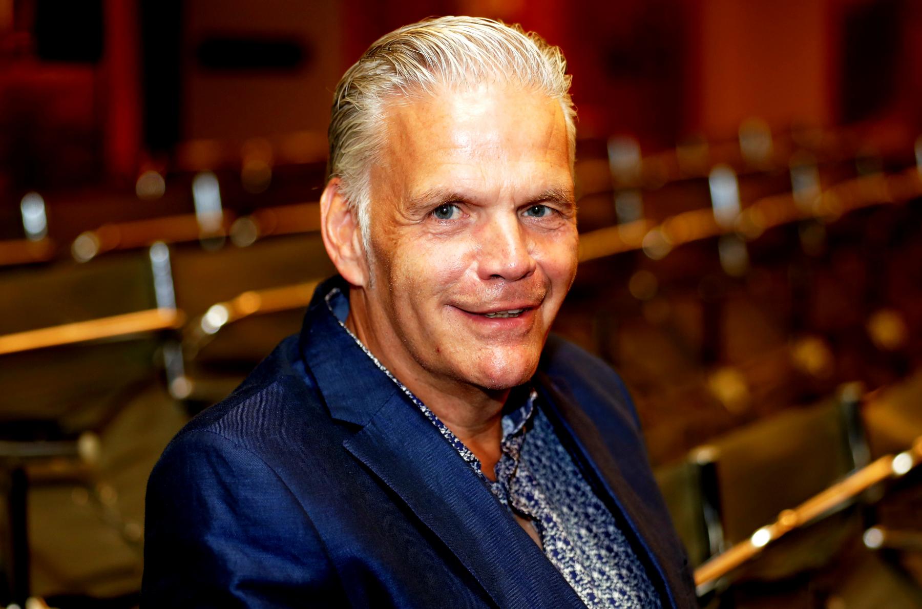 Portrait eines älteren Mannes mit weißen, kurzen Haaren im Zuschauerraum eines Theaters. Er trägt ein blaues Sakko über einem blau-weiß gemusterten Hemd und lacht freundlich in die Kamera.