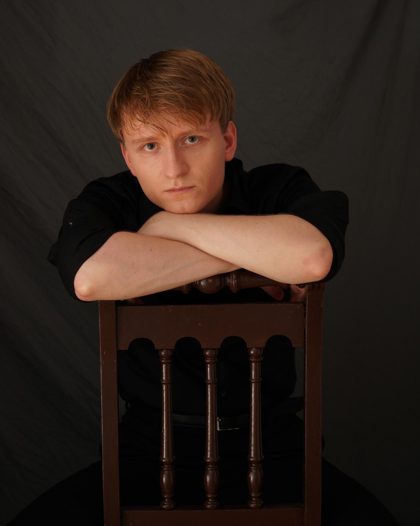 Portrait von Nino Suska, ein junger Mann der vor einem dunklen Hintergrund auf einem Stuhl sitzt und sich auf der Rückenlehne abstützt.