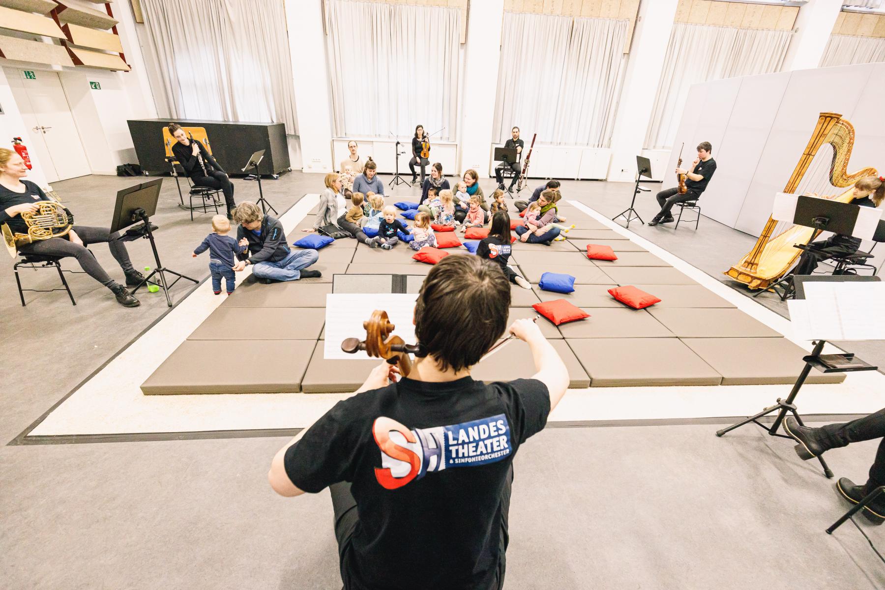 Kleinkinder und ihre Eltern tummeln sich auf dem Boden. Um sie herum sitzen Musiker und muszieren.