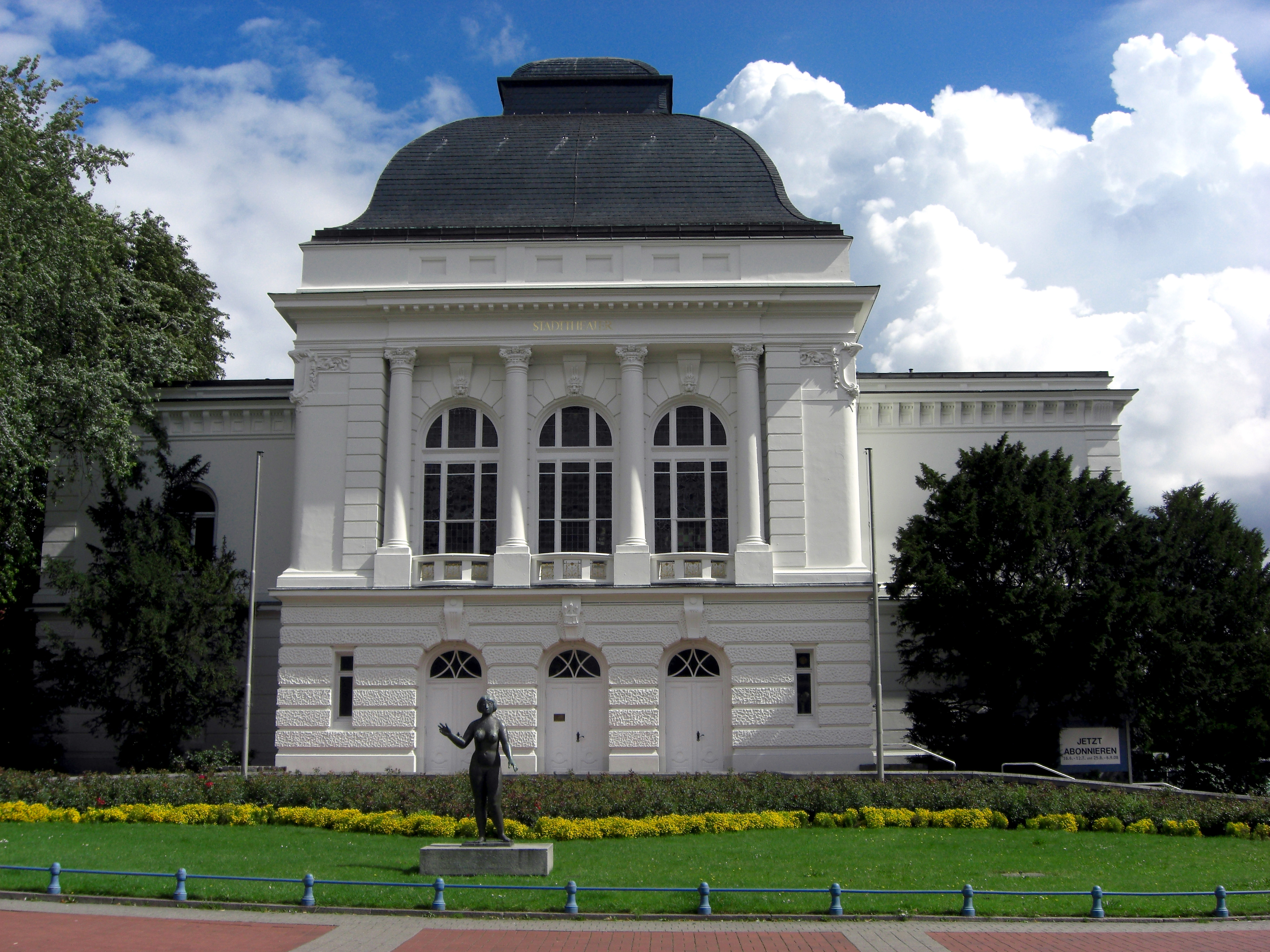 Bild des Stadttheaters Rendsburg von außen, ein großes weißes Gebäude mit weißen Säulen, großen Fenstern und einem dunklem Dach, davor eine grüne Rasenfläche mit Blumen