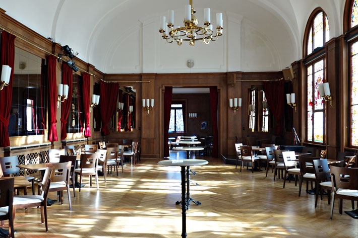 Das Große Foyer im Stadttheater Rendsburg, an den Seiten des imposanten Raumes stehen Tische und Stühle, der Raum ist mit dunklem Holz verkleidet, an der rechten Wand sind große, bunte Fenster zu sehen