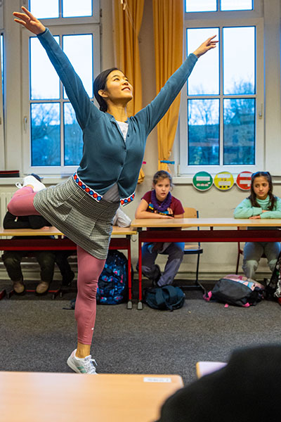 Kinder in einem Klassenzimmer, in der Mitte des Raumes tanzt eine junge Frau in bunter Kleidung