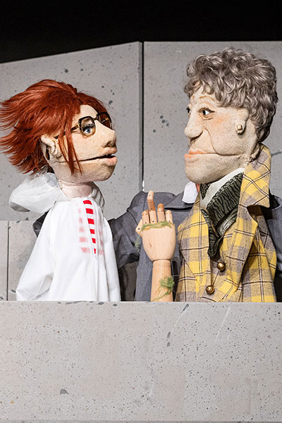 Zwei Puppen schauen einander zwischen grauen Betonwänden an, die linke Puppe ist rothaarig, trägt eine Brille und einen weißen Kittel, die rechte Puppe hat kurze graue Haare, trägt einen gelb-karierten Mantel, zwischen ihnen eine hölzerne Hand mit Algen daran