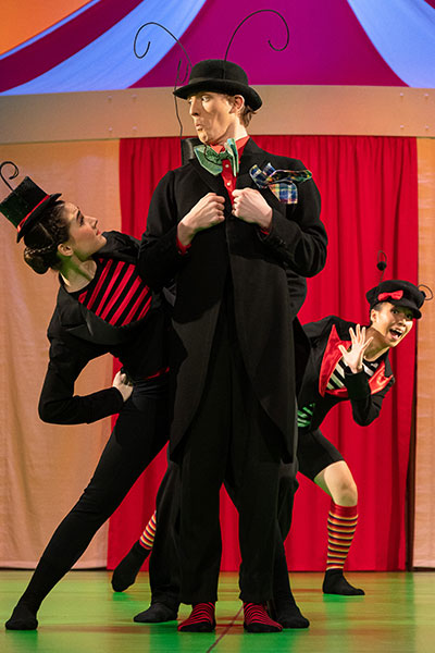Drei Personen in schwarz-roten Anzügen stehen auf der Bühne, im Hintergrund ein Zirkuszelt, sie stehen hintereinander und schauen an den Seiten hervor, sie blicken einander verspielt an