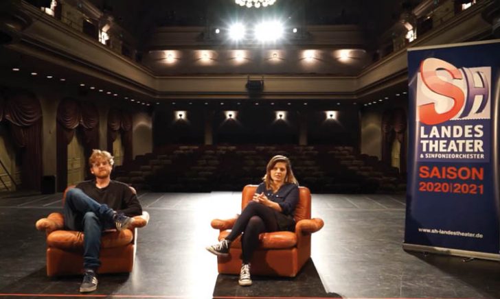 Blick von der Bühne in den leeren Zuschauerraum, auf der Bühne sitzen zwei Personen in gemütlichen Ledersesseln und schauen zur Kamera, neben ihnen steht ein Banner mit dem Logo des Landestheaters und der Aufschrift "SAISON 2020/2021"