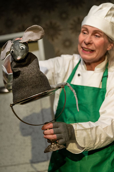 Eine Frau ist gekleidet als Koch, mit weißer Mütze und grüner Schürze, sie hält eine Puppe in der Form einer Maus vor sich und lächelt diese an