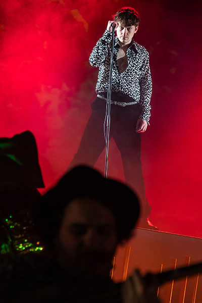 Ein junger Mann steht allein auf einer rot erleuchteten Bühne, im Hintergrund ist Nebel, er hält ein Mikrofon auf einem Ständer in der Hand und blickt ernst nach vorn