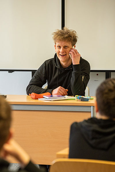 In einem Klassenzimmer sitzt ein junger Mann mit wirrem Haar am Pult und telefoniert, dabei lacht er