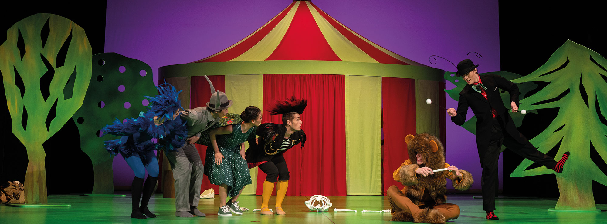 Szene aus DER KARNEVAL DER TIERE, Zirkuslandschaft, ein Löwe hält einen Kochen in der Hand, um ihn herum weitere Tiere