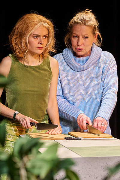 Zwei Frauen, die mit Messern und Schneidebrettern an einer erhöhten Oberfläche stehen, die junge Frau links schaut ernst in die Ferne, die etwas ältere Frau rechts spricht mit ihr.