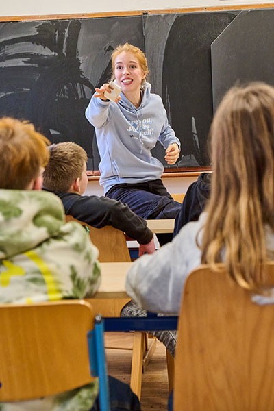 Ein Klassenzimmer voll mit jungen Schülerinnen und Schülern, eine junge Frau in einem hellblauen Hoodie sitzt auf dem Pult, sie streckt ihren Arm nach vorne aus, in der Hand hält sie ein Smartphone