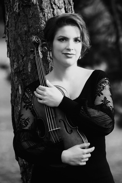 Schwarz-weiß Dreiviertelportrait einer jungen Frau mit kurzen, dunklen Haaren und im schwarzen Abendkleid mit durchsichtigen, bestickten langen Ärmeln. Im rechten Arm hält sie ihre Violine. Sie leht an einem Baumstamm.