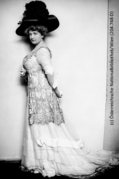Schwarz-weiß Ganzkörperportrait von Alma Mahler-Werfel, sie steht zur Seite gedreht mit dem Blick zur Kamera, die Hänge hinter dem Rücken gekreuzt, in einem eleganten bodenlangen Kleid und einem federbesetzten enormen Hut