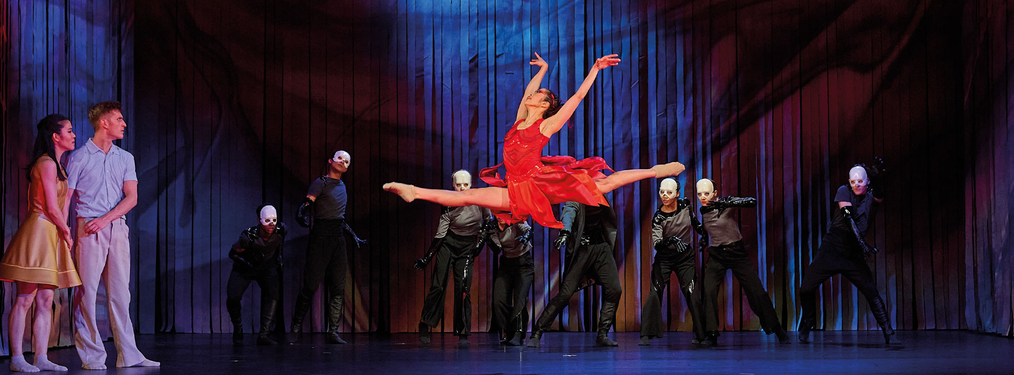 Aus DER FEUERVOGEL, Tänzerin in rotem Kleid, hinter ihr dunkle Gestalten, ein Paar steht links im Bild